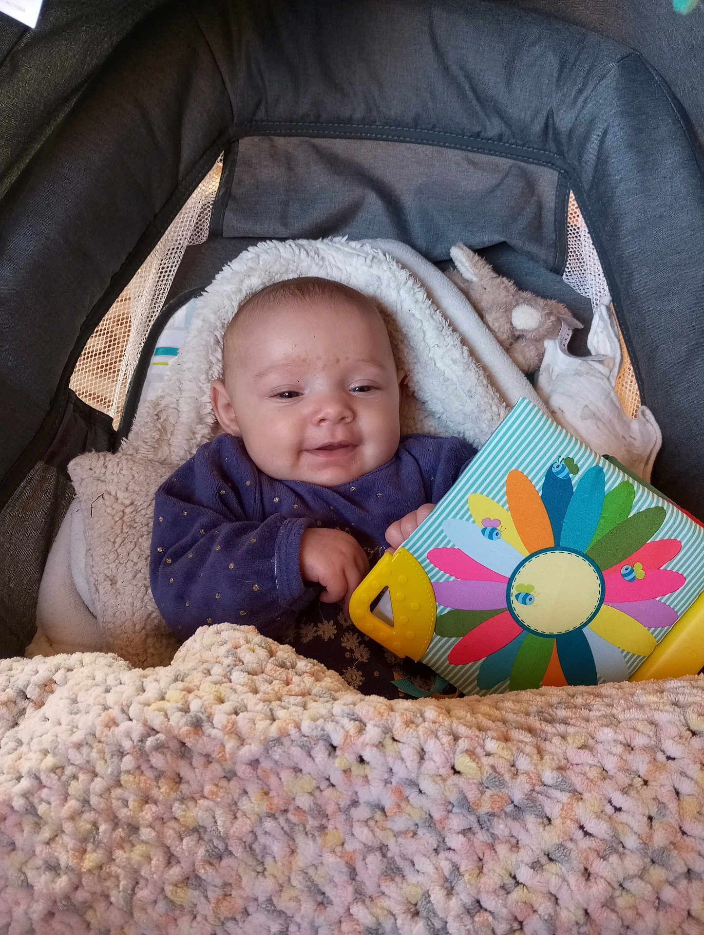 Clara participe au concours pour gagner de l'argent avec cette photo : baby, infant, child, stroller, blanket, knit_blanket, toy_book, colorful_toy, smile, happy, face, hand, purple_clothing, plush_toy, bedding, cozy, portrait, indoor, closeup, young