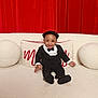 toddler, child, smiling, suit, bow_tie, hat, couch, cushion, red_curtain, indoor, portrait, happy, seated, fashion, formal_wear, baby, person, cute, cozy, photography