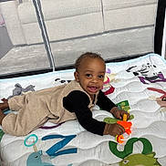 Denorio is registered to the contest to win money with this photo: baby, child, playpen, play_mat, toy, smiling, indoor, couch, mesh, clothing, person, happy, lying_down, infant, colorful, black_shirt, beige_outfit, bright_toy, curious, young_child