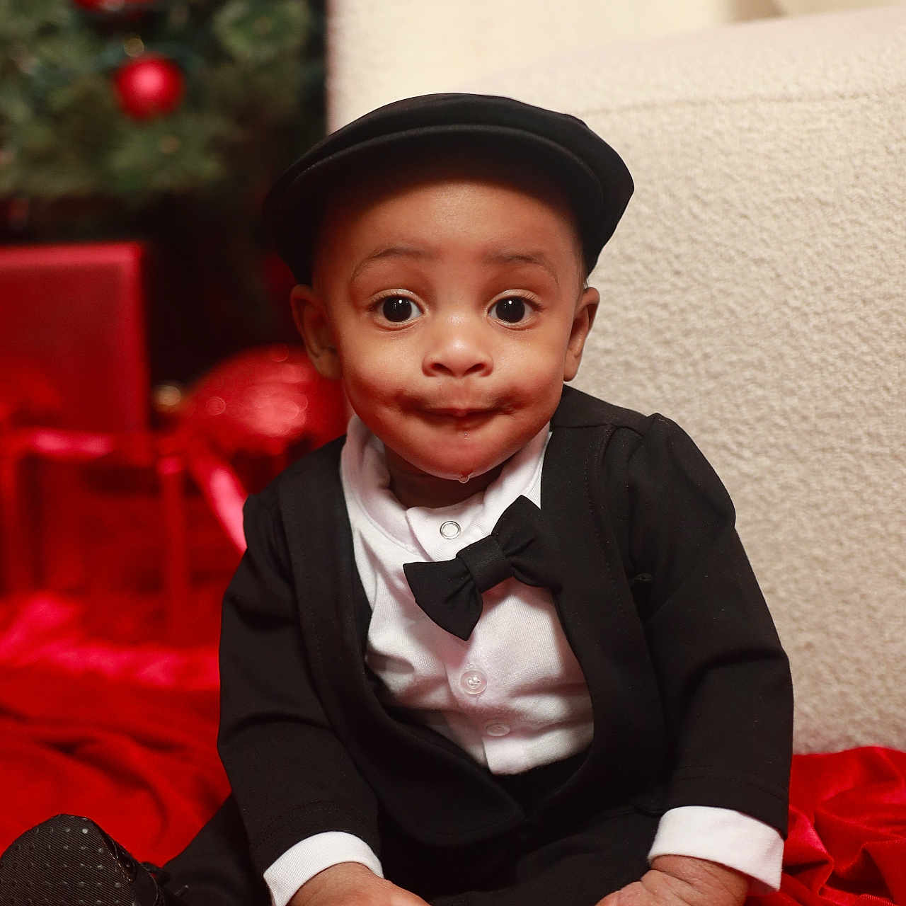 Denorio is registered to the contest to win money with this photo: baby, black_clothing, bow_tie, child, christmas_decorations, christmas_tree, cute, expression, face, hands, hat, holiday, indoors, portrait, presents, red_blanket, sitting, toddler, tuxedo, white_shirt