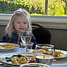 child, girl, blonde_hair, pink_hair_clip, denim_jacket, dining_table, plate, food, glass, water, window, greenery, chair, indoor, meal, fork, napkin, light, smile, person