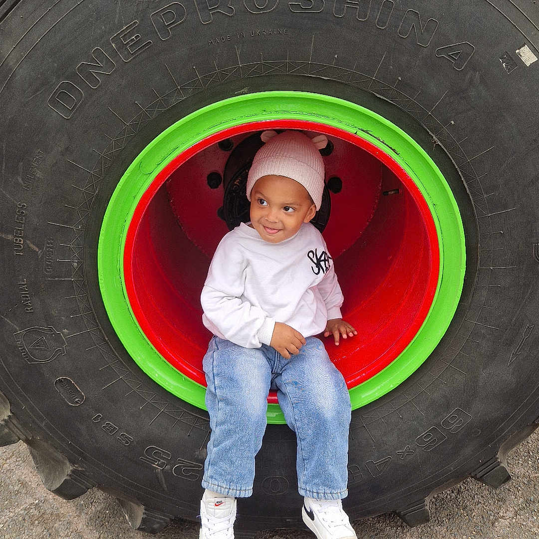 Soan a rejoint le concours — aidez-le/la à gagner de superbes lots ! beanie, blue_jeans, casual_clothing, child, concrete_ground, cute, daylight, fun, green, outdoor, playful, red, sitting, smiling, tire_rim, toddler, tractor_tire, white_sneakers, white_sweatshirt, young_child
