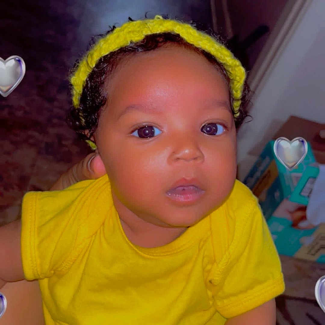 Lainey is registered to the contest to win money with this photo: baby, child, yellow_clothing, headband, indoor, person, cute, infant, skin, hand, face, eyes, curious, holding, human, portrait, young_child, heart_overlay, bright_color, floor