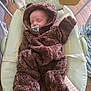 baby, sleeping, pacifier, brown_onesie, hood, baby_rocker, frog_design, wooden_floor, toy, infant, cozy, cute, child, soft_clothing, indoor, resting, comfort, small_face, peaceful, wearing