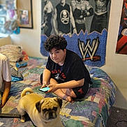 Max is registered to the contest to win money with this photo: person, dog, pug, bed, phone, poster, wwe, room, indoor, carpet, curly_hair, black_shirt, shorts, furniture, blanket, remote_control, wall_art, relaxed, casual, sitting