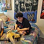 person, dog, pug, bed, phone, poster, wwe, room, indoor, carpet, curly_hair, black_shirt, shorts, furniture, blanket, remote_control, wall_art, relaxed, casual, sitting