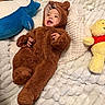 baby, child, plush_toy, stuffed_animal, brown_onesie, bear_costume, smiling, cozy_blanket, white_blanket, blue_plush, yellow_toy, red_shirt, infant, cute, happy, lying_down, soft_texture, indoors, portrait, warm