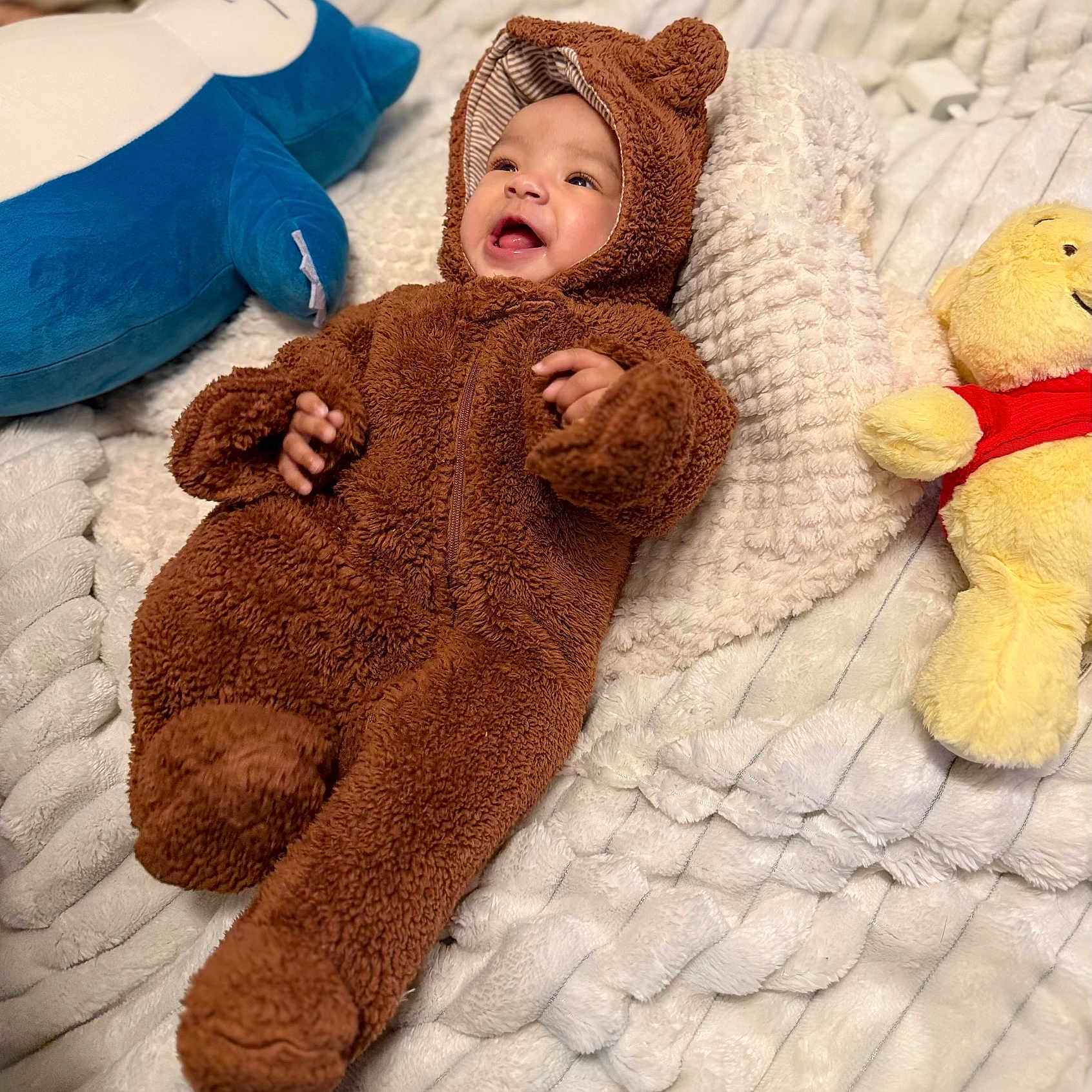 Aavani joined the competition — help win amazing prizes! baby, child, plush_toy, stuffed_animal, brown_onesie, bear_costume, smiling, cozy_blanket, white_blanket, blue_plush, yellow_toy, red_shirt, infant, cute, happy, lying_down, soft_texture, indoors, portrait, warm