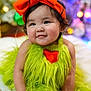 Myah joined the competition — help win amazing prizes! baby, child, infant, headband, orange_bow, green_outfit, fuzzy_clothing, heart_patch, smiling, cute, portrait, indoors, colorful_background, holiday_lights, festive, soft_lighting, happy, sitting, closeup, adorable