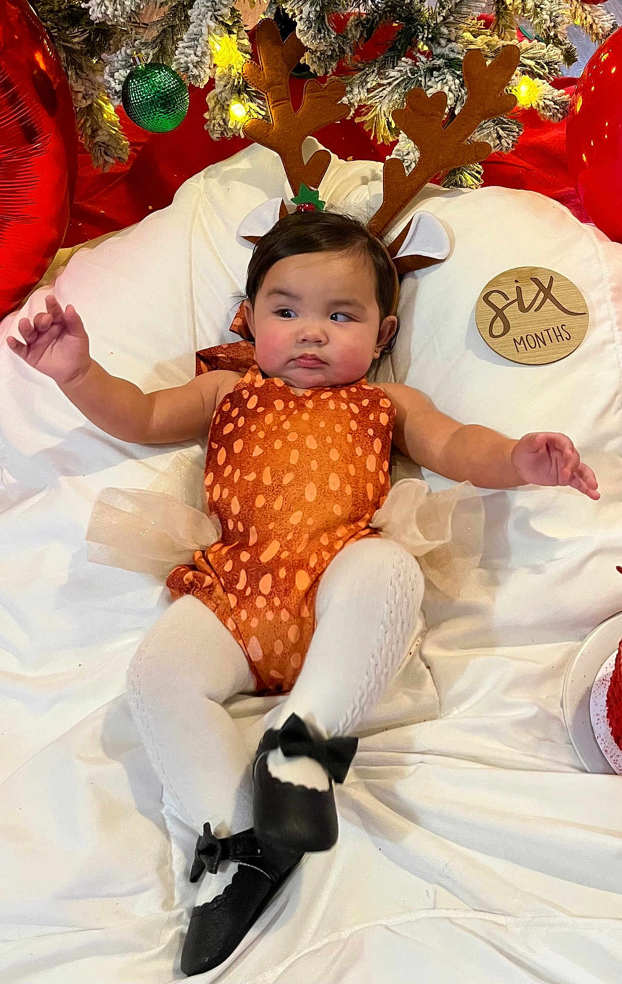 Myah is registered to the contest to win money with this photo: baby, child, reindeer_costume, antlers, christmas, holiday, festive, decorations, green_ornament, red_balloons, white_tights, black_shoes, plush_cushion, six_months, wooden_plaque, cute, infant, portrait, indoors, celebration