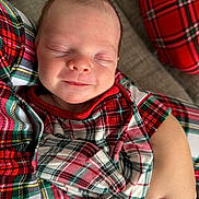 Elizabeth is registered to the contest to win money with this photo: baby, newborn, infant, sleeping, smiling, face, cheek, head, plaid, pajamas, pattern, red, green, blanket, couch, arm, hand, close_up, portrait, cozy