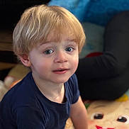 Ezekiel is registered to the contest to win money with this photo: child, toddler, blonde_hair, blue_eyes, indoor, play_mat, cartoon, blanket, person, clothing, curious, sitting, face, portrait, young_child, casual, home, expression, people, background