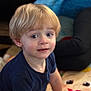 child, toddler, blonde_hair, blue_eyes, indoor, play_mat, cartoon, blanket, person, clothing, curious, sitting, face, portrait, young_child, casual, home, expression, people, background