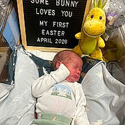 Ma'Kai is registered to the contest to win money with this photo: baby, newborn, sleeping, pajamas, plush_toy, yellow_toy, letter_board, text_message, blanket, bed, indoor, cute, infant, resting, child, soft_toy, announcement, easter, celebration, portrait