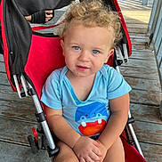 Benjamin is registered to the contest to win money with this photo: child, toddler, curly_hair, blue_eyes, stroller, red_stroller, blue_shirt, cartoon_hippo, porch, wooden_floor, hands_clasped, outdoor, sunlight, casual_clothing, baby, person, seated, cute, expression, curious