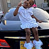 A'Moura is registered to the contest to win money with this photo: child, girl, smiling, sitting, car, black_lexus, white_dress, colorful_beads, hair_braids, shoes, outdoor, parking_lot, apartment_building, license_plate, happy, playful, pose, finger_gun, daylight, urban