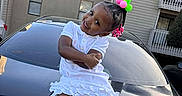 A'Moura is registered to the contest to win money with this photo: toddler, girl, child, car, lexus, hair_accessories, white_dress, playful_pose, outdoor, apartment_building, smiling, crossed_arms, daytime, shoes, license_plate, black_car, vehicle, cute, fun, portrait