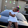 toddler, girl, child, car, lexus, hair_accessories, white_dress, playful_pose, outdoor, apartment_building, smiling, crossed_arms, daytime, shoes, license_plate, black_car, vehicle, cute, fun, portrait