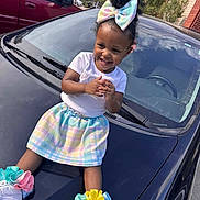 A'Moura is registered to the contest to win money with this photo: accessories, bright_colors, car_hood, casual_clothing, child, clapping, colorful_bow, curly_hair, daylight, happy, outdoor, pastel_skirt, playful, residential_area, smiling, sneakers, toddler, trees, vehicle, white_tshirt