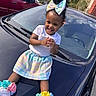 accessories, bright_colors, car_hood, casual_clothing, child, clapping, colorful_bow, curly_hair, daylight, happy, outdoor, pastel_skirt, playful, residential_area, smiling, sneakers, toddler, trees, vehicle, white_tshirt