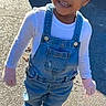 child, toddler, smiling, outdoor, sunlight, denim_overalls, blue_bow, white_shirt, sneakers, pavement, shadow, happy, person, standing, cute, young_child, casual_clothing, daylight, playful, small_child