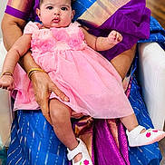 Tara joined the competition — help win amazing prizes! baby, child, pink_dress, white_shoes, flower_design, adult_hand, traditional_clothing, purple_saree, blue_saree, seated, indoor, person, cute, portrait, infant, looking_at_camera, holding, hand_bracelet, gold_jewelry, fabric