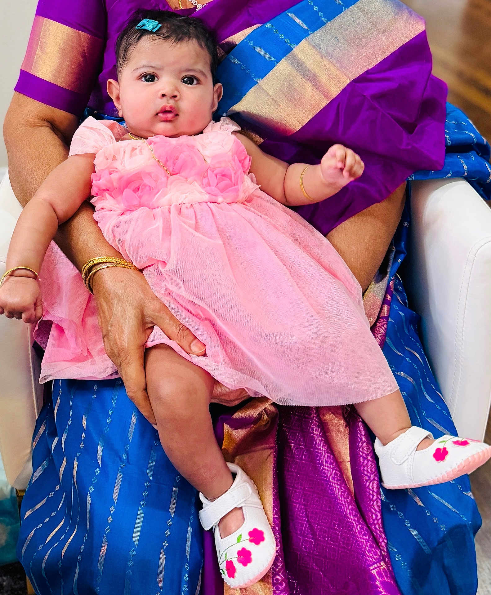Tara joined the competition — help win amazing prizes! baby, child, pink_dress, white_shoes, flower_design, adult_hand, traditional_clothing, purple_saree, blue_saree, seated, indoor, person, cute, portrait, infant, looking_at_camera, holding, hand_bracelet, gold_jewelry, fabric