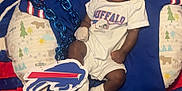 Ethan is registered to the contest to win money with this photo: baby, sleeping, onesie, buffalo, football, sports_fan, blanket, hat, chain, logo, pillow, blue, red, white, infant, cute, cozy, indoor, person, child