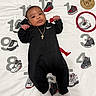 baby, infant, black_clothing, nike, gold_chain, blanket, sneakers, numbers, two_months, wooden_plaque, red_circle, cute, lying_down, milestone, portrait, child, indoors, white_blanket, cartoon_shoes, curious