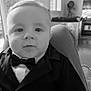 baby, black_and_white, bow_tie, child, close_up, clothing, cute, face, formal_wear, furniture, home, indoor, infant, person, portrait, sitting, smile, tuxedo, window, young_child