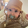 baby, baby_boy, baby_girl, blue_eyes, brown_hat, child, clothing, curious, cute, face, hat, high_chair, indoor, infant, long_eyelashes, overalls, pacifier, person, seated, soft_light