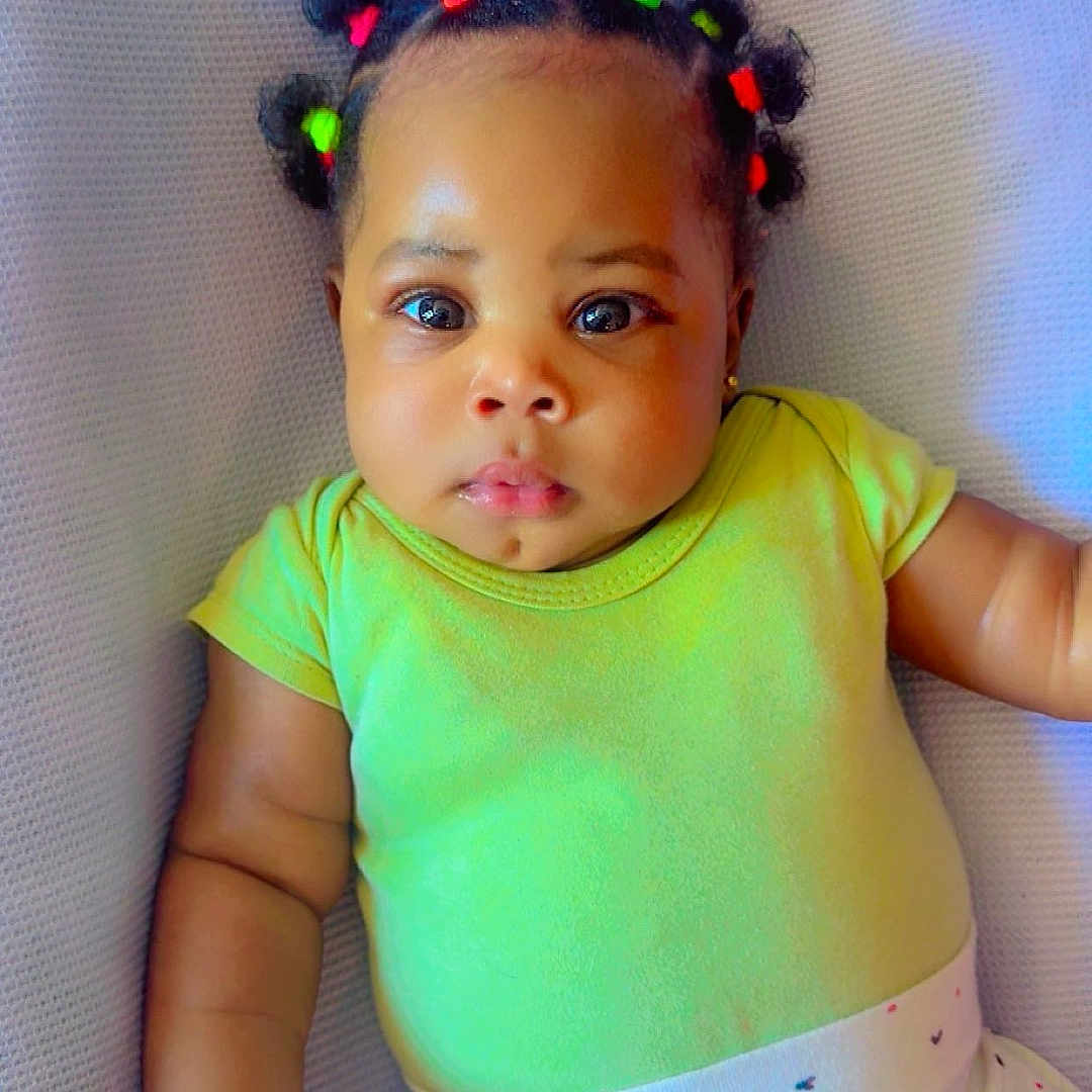 Athena-Amaris is registered to the contest to win money with this photo: adorable, baby, big_eyes, child, colorful, curious_expression, cushion, cute, face, green_onesie, hair_ties, hand, indoor, infant, lying_down, patterned_clothing, portrait, skin, soft_texture, white_pants