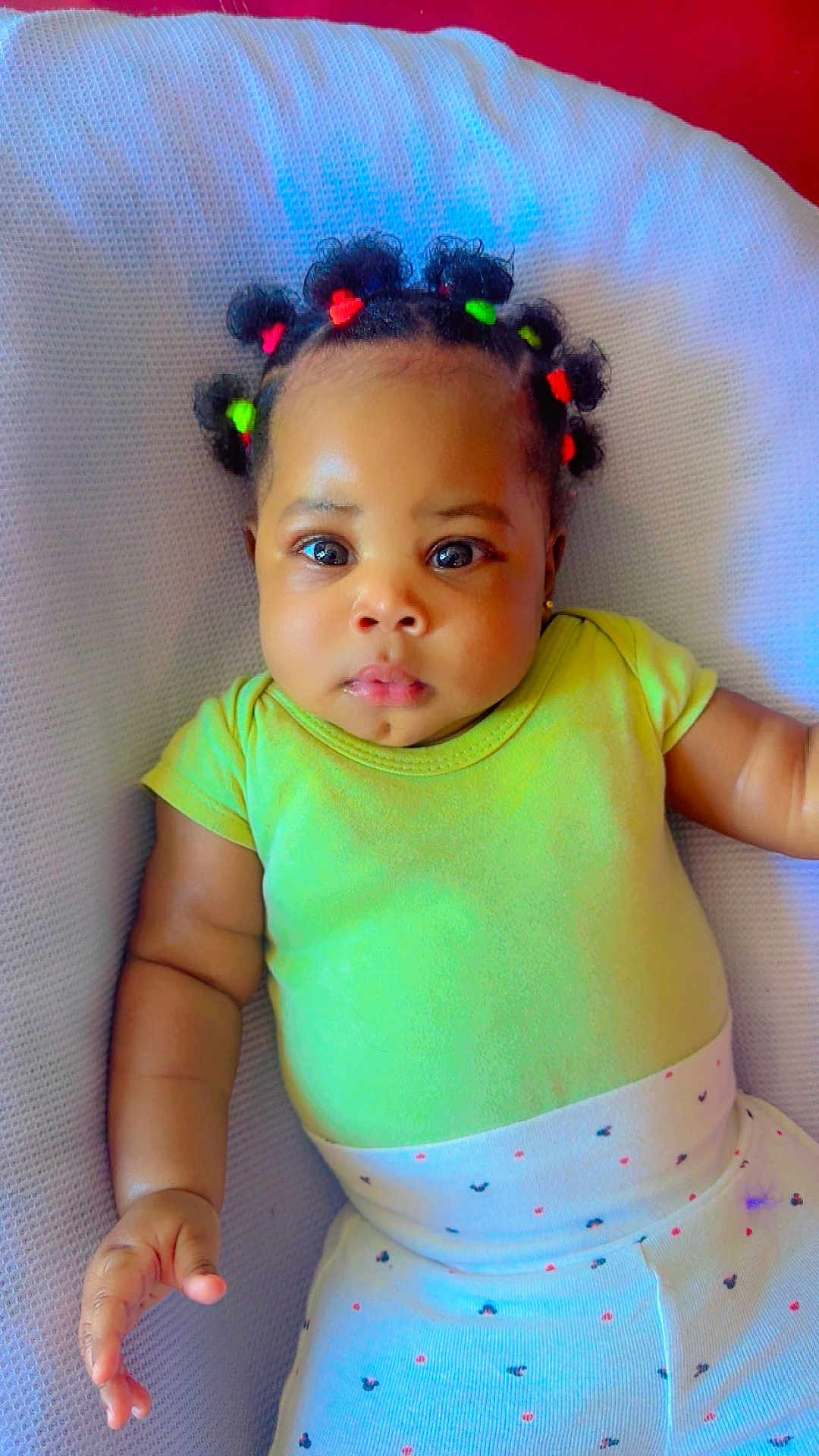Athena-Amaris is registered to the contest to win money with this photo: baby, child, infant, cute, hair_ties, colorful, green_onesie, white_pants, patterned_clothing, cushion, soft_texture, lying_down, curious_expression, big_eyes, indoor, portrait, skin, hand, face, adorable