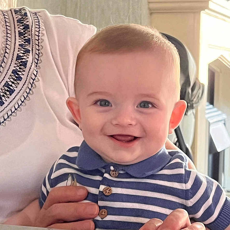 Eoghan is registered to the contest to win money with this photo: adult_hand, baby, bright, child, cute, face, hand, happy, holding, home, indoor, infant, light, person, polo_shirt, portrait, smiling, striped_shirt, table, young_child