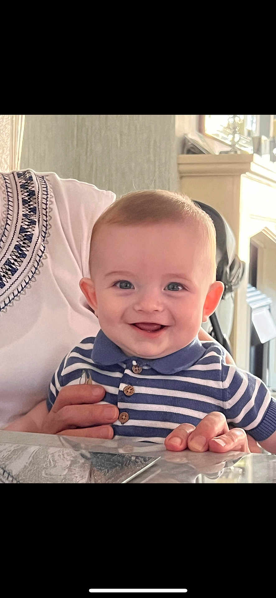 Eoghan is registered to the contest to win money with this photo: baby, child, smiling, happy, face, person, striped_shirt, polo_shirt, hand, adult_hand, indoor, home, table, portrait, cute, young_child, infant, holding, bright, light