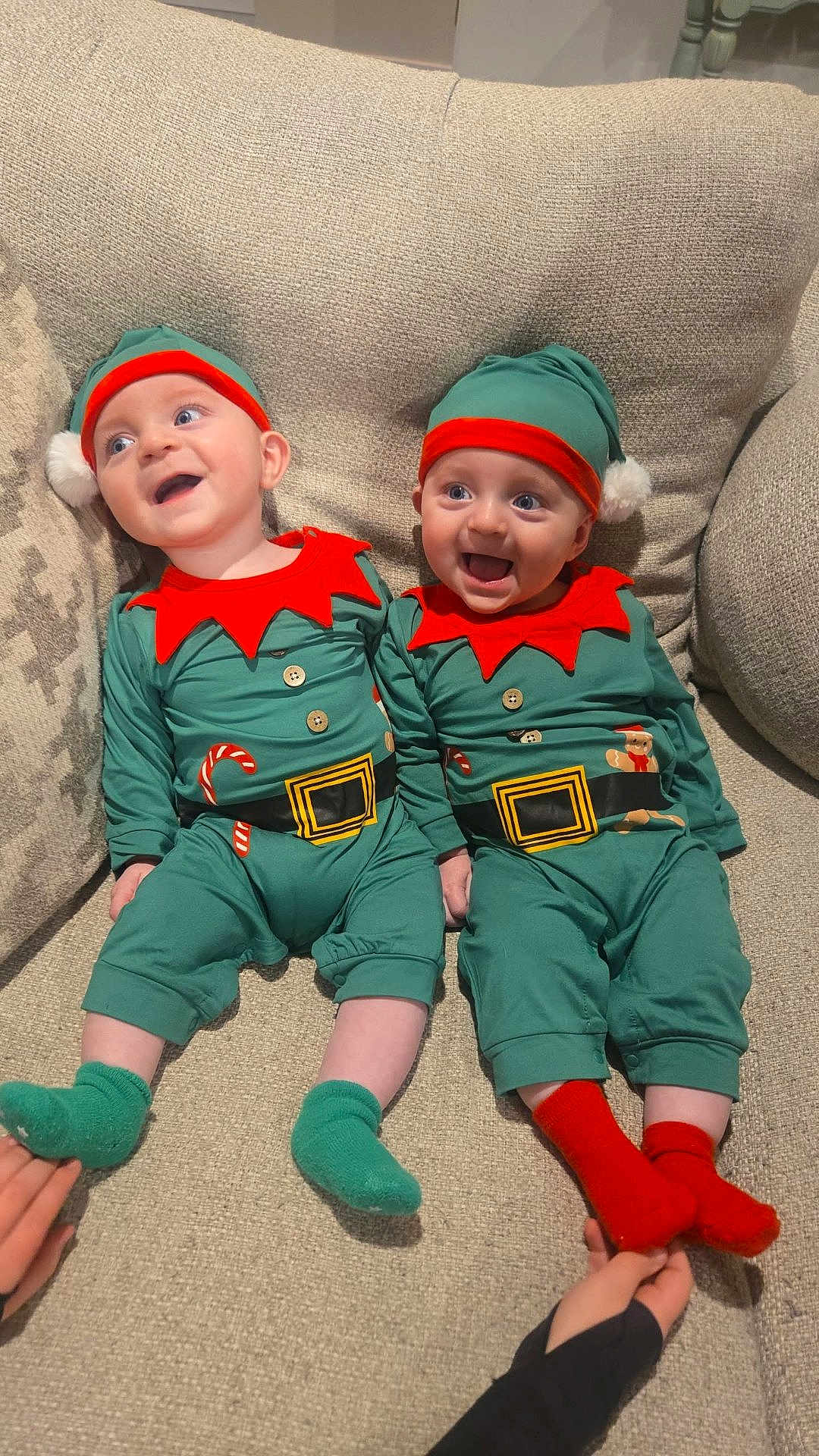 Collin And Kyler is registered to the contest to win money with this photo: baby, infant, toddler, child, elf_costume, holiday_outfit, green_hat, red_trim, couch, beige_couch, smiling, happy, cute, festive, holiday, socks, green_socks, red_socks, two_babies, indoor