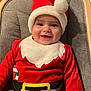 baby, infant, smiling, santa_hat, christmas_outfit, red_clothing, white_trim, baby_chair, cushion, wooden_frame, happy, face, child, holiday, festive, indoors, seat_belt, portrait, cute, celebration