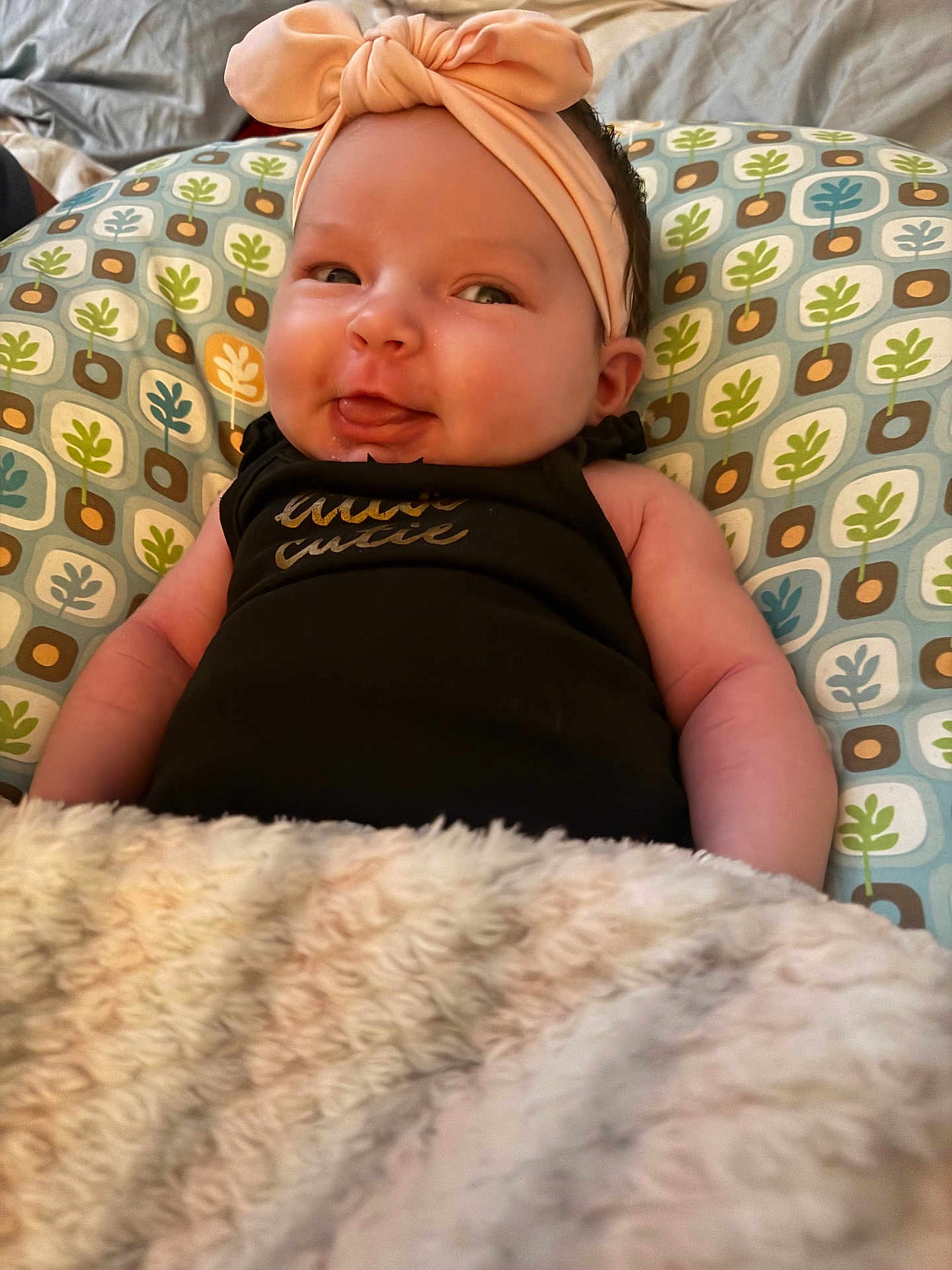 Ariya is registered to the contest to win money with this photo: baby, infant, headband, blanket, patterned_pillow, smiling, tongue_out, cute, cozy, child, soft_texture, indoors, clothing, human, resting, face, cheeks, expression, portrait, relaxing