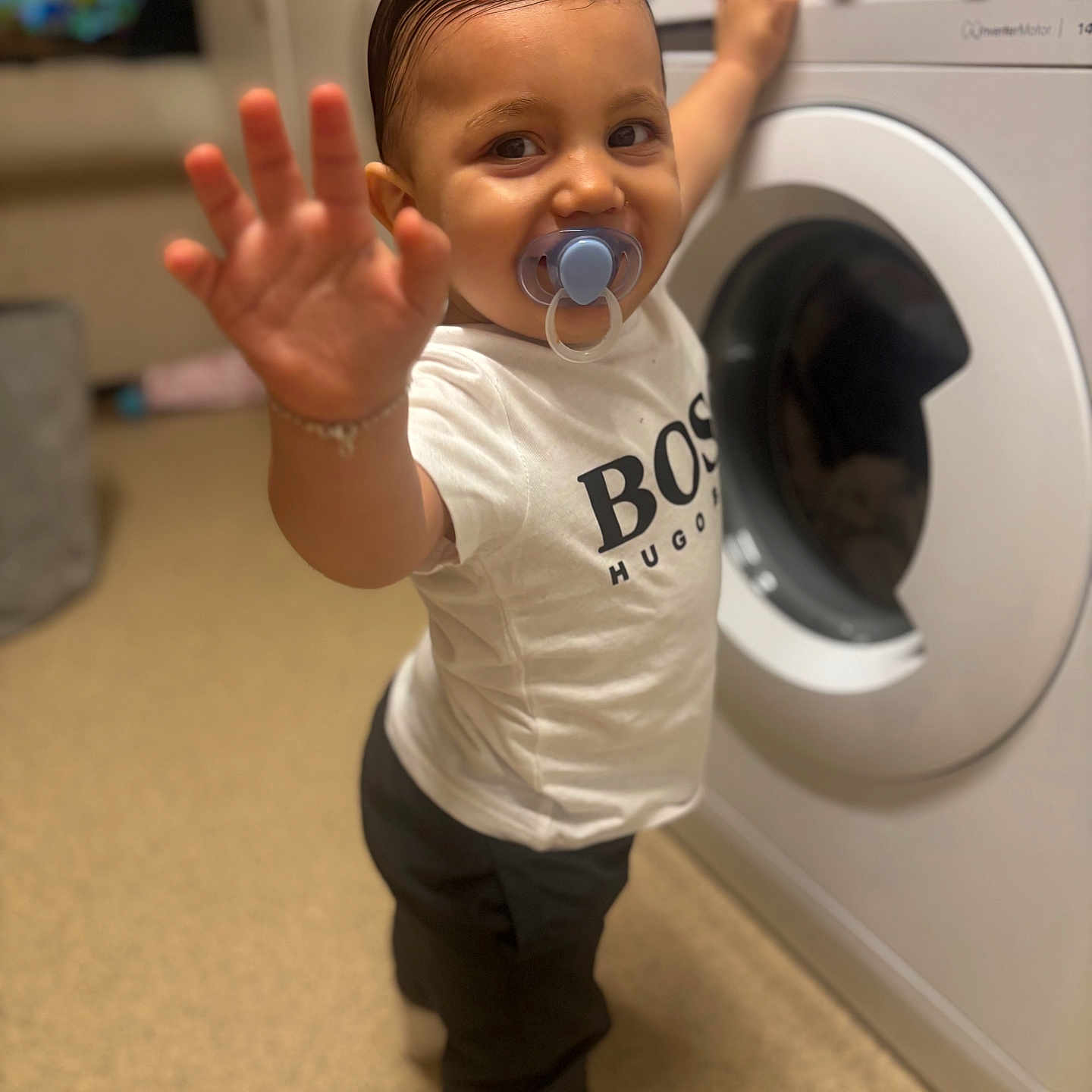 Teddie Lee joined the competition — help win amazing prizes! appliance, black_pants, child, clothing, curious, flooring, hair, hand, home, household, indoor, pacifier, person, playful, smiling, standing, toddler, washing_machine, white_tshirt, young_child