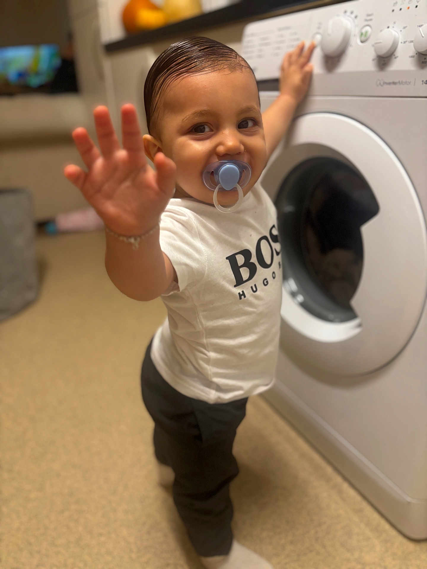Teddie Lee joined the competition — help win amazing prizes! toddler, child, pacifier, washing_machine, white_tshirt, black_pants, indoor, home, flooring, smiling, hand, person, young_child, clothing, hair, appliance, playful, standing, household, curious