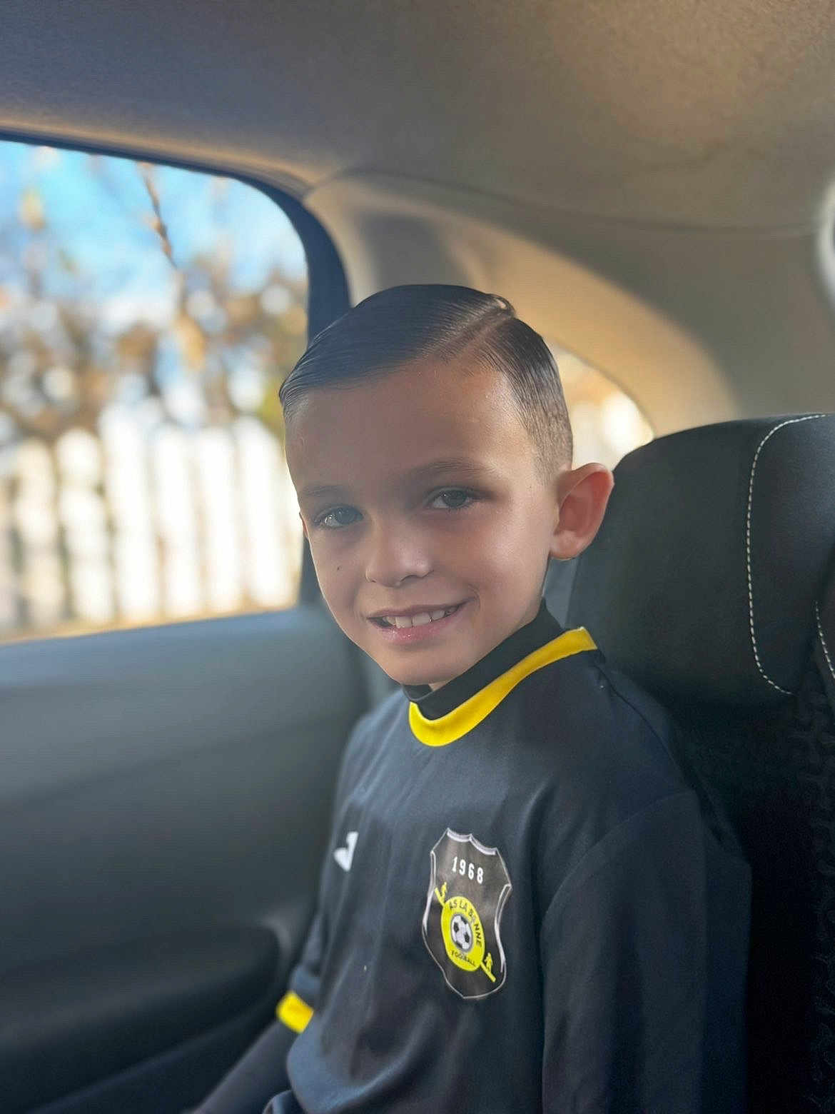 Aarôn a rejoint le concours — aidez-le/la à gagner de superbes lots ! boy, child, smile, car, window, sports_jersey, black_clothing, yellow_trim, hairstyle, seat, seatbelt, interior, portrait, young, casual, daylight, background_blur, happy, person, sitting