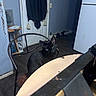 black_cat, cat, chair, table, indoor, kitchen, refrigerator, door, curtain, wooden_floor, side_table, papers, metal_chair, yellow_eyes, pet, domestic_animal, furniture, home_interior, floor, wall