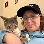 Pip is registered to the contest to win money with this photo: person, cat, glasses, cap, hoodie, indoor, smile, pet, animal, face, hand, furniture, wall, picture_frame, cozy, casual, domestic, cute, holding, portrait