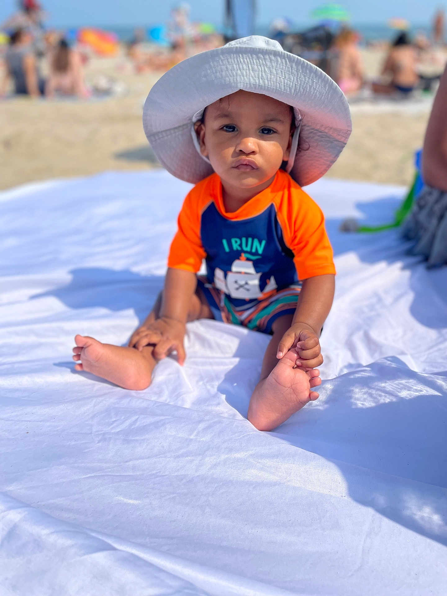 Ni Kai joined the competition — help win amazing prizes! toddler, child, beach, hat, sunhat, orange_clothing, blue_clothing, blanket, sand, people, umbrella, summer, outdoor, sunny, portrait, baby, feet, sitting, vacation, casual