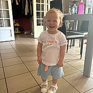 Julia a rejoint le concours — aidez-le/la à gagner de superbes lots ! toddler, child, smiling, standing, indoor, tile_floor, white_tshirt, blue_skirt, white_shoes, furniture, chair, table, lamp, bottle, water_bottle, vacuum_cleaner, door, clothes, happy, person