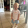 toddler, child, smiling, standing, indoor, tile_floor, white_tshirt, blue_skirt, white_shoes, furniture, chair, table, lamp, bottle, water_bottle, vacuum_cleaner, door, clothes, happy, person