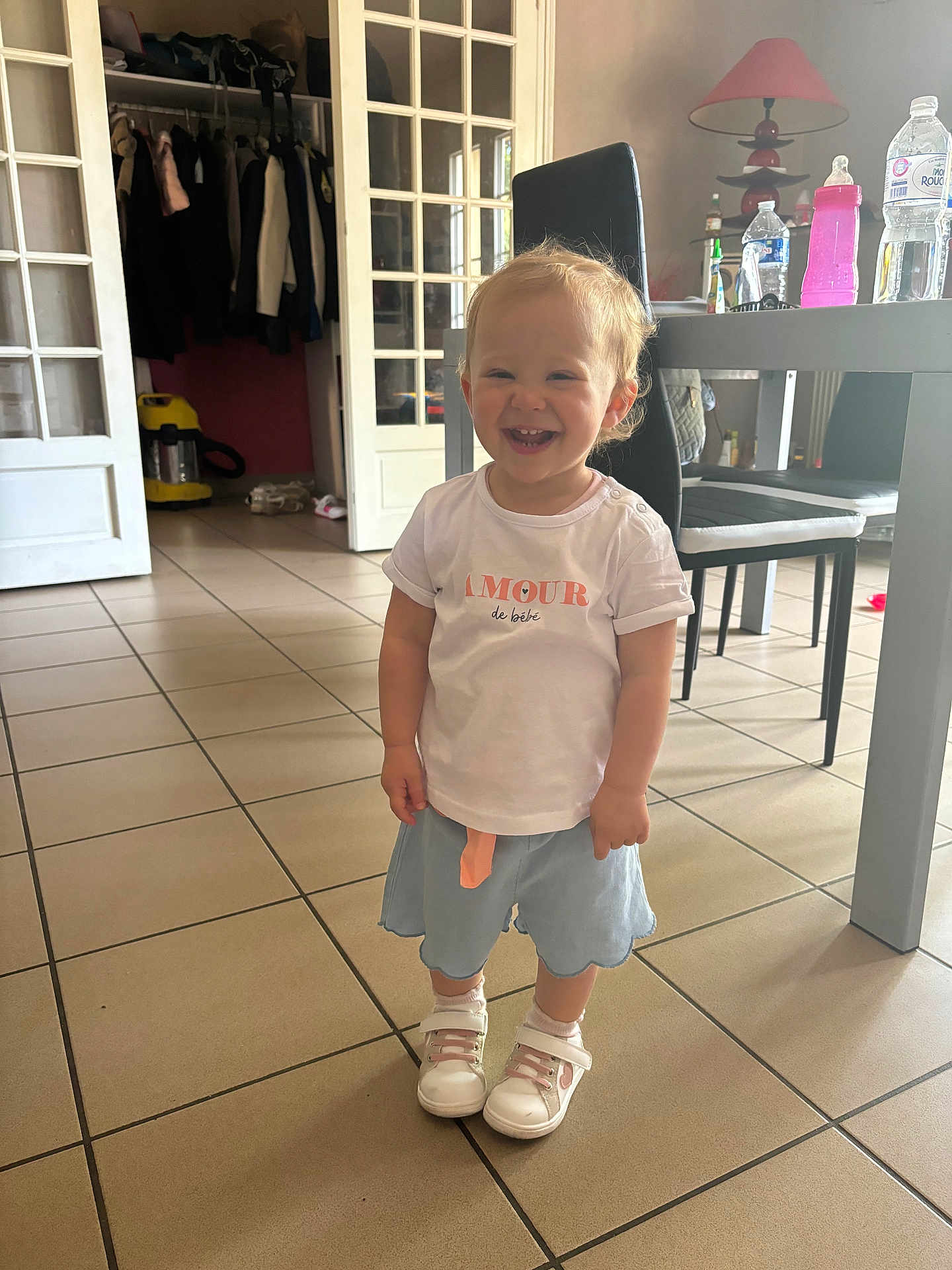 Julia a rejoint le concours — aidez-le/la à gagner de superbes lots ! toddler, child, smiling, standing, indoor, tile_floor, white_tshirt, blue_skirt, white_shoes, furniture, chair, table, lamp, bottle, water_bottle, vacuum_cleaner, door, clothes, happy, person
