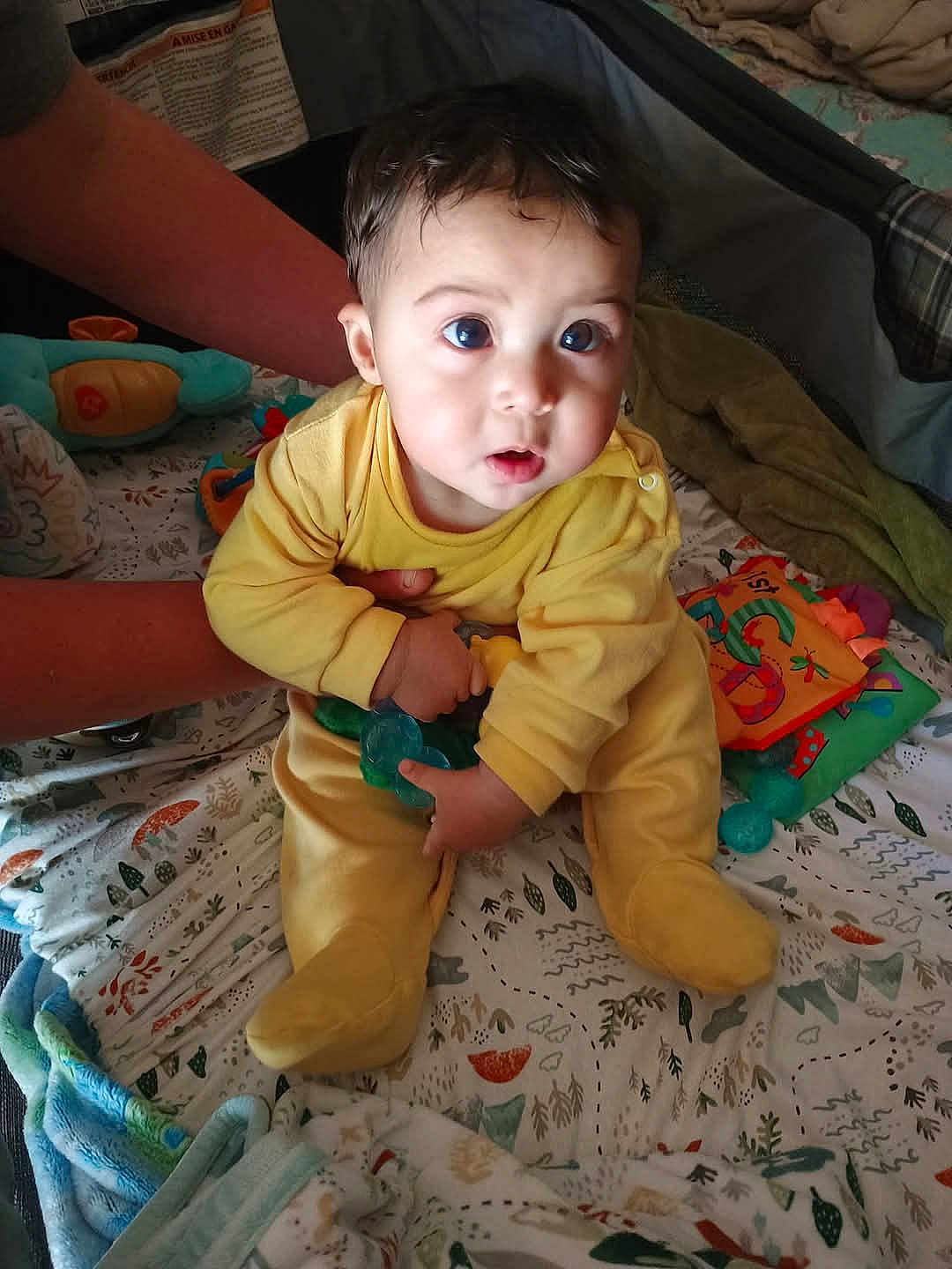 Jaxton joined the competition — help win amazing prizes! baby, child, yellow_onesie, toy, blanket, hands, indoor, cute, curious, infant, person, play_area, soft_toys, patterned_blanket, supporting_hands, sitting, wide_eyes, cozy, close_up, nursery