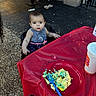 cake, celebration, child, clothing, concrete_floor, curious, daylight, food, footwear, fork, high_chair, outdoor, party, person, picnic_table, plastic_cup, plate, red_tablecloth, seat, toddler