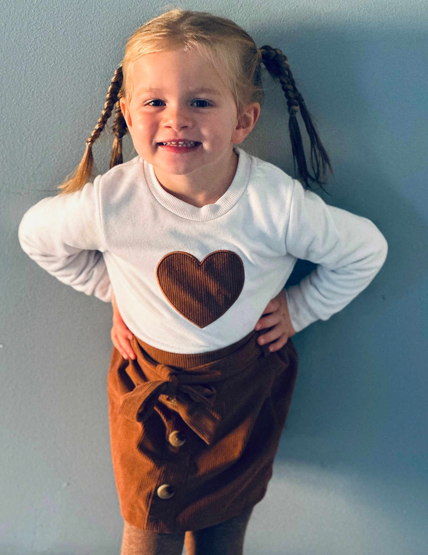 Iris a rejoint le concours — aidez-le/la à gagner de superbes lots ! child, girl, smiling, braids, pigtails, white_sweater, heart, brown_skirt, buttons, standing, posing, indoor, wall, happy, cute, fashion, portrait, young, person, clothing